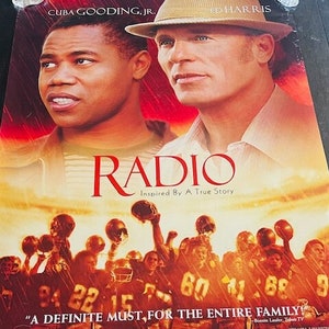 Può includere: Locandina del film "Radio", ispirato a una storia vera, con Cuba Gooding Jr. e Ed Harris. La locandina presenta una combinazione di colori rosso e arancione con un gruppo di giocatori di football sullo sfondo. Il testo "Un must assoluto per tutta la famiglia" è presente nella parte inferiore della locandina.