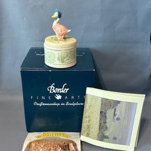 Puede incluir: Una figurita de porcelana de Jemima Puddle-Duck de El cuento de Peter Rabbit de Beatrix Potter, con un vestido rosa y un gorro azul, se sienta encima de una caja de música redonda de cerámica blanca con un diseño floral verde y blanco. La caja de música está en su caja original, que es azul con letras doradas que dicen "Border Fine Arts Craftsmanship in Sculpture".