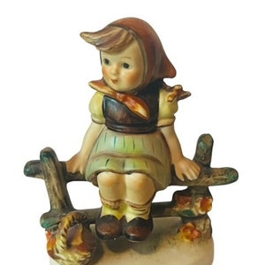 Op de afbeelding: Een porseleinen beeldje van een meisje in een rood-bruine jurk dat op een houten hek zit met een eekhoorn aan haar voeten.