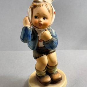 May include: A vintage porcelain figurine of a young child wearing a white headscarf with red polka dots, a blue plaid jacket, and brown shorts. The figurine is standing on a round, beige base. The child's expression is one of surprise or concern.