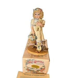Könnte beinhalten: Eine Porzellanfigur eines jungen Mädchens, das einen Teddybär hält. Sie trägt ein weißes Kleid mit einem blauen und rosa Band im Haar. Sie steht auf einem Sockel mit zwei Teddybären. Die Figur befindet sich in einer Schachtel mit den Worten "Jan Wabara" und "Limitierte Auflage".