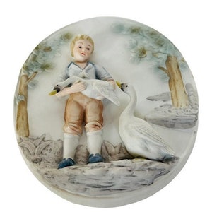 May include: A porcelain figurine of a young person holding two white swans, with a third swan nearby. The figurine is set against a backdrop of trees and foliage, in a circular shape. The figure is wearing a blue shirt, brown trousers, and blue shoes.