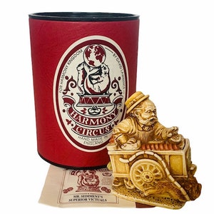 May include: A hand-painted ceramic figurine of a man with a beard, wearing a hat and sitting on a cart with a sign that reads "Harmony Circus". The cart is decorated with a globe and the words "Hand Made in England".