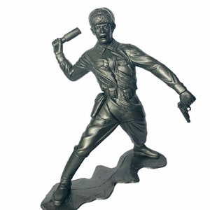 Peut inclure: Une figurine de soldat en plastique gris portant un chapeau et tenant un fusil dans une main et une grenade dans l'autre. Le soldat est debout sur une base en plastique gris.