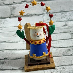 Könnte beinhalten: Ein skurriles Ornament mit einem Marshmallow-Charakter mit lächelndem Gesicht, der ein blaues Outfit mit gelben Verzierungen und roten Herzakzenten trägt. Das Ornament hält eine Girlande aus Sternen und Herzen und steht auf einer Graham-Cracker-Basis.