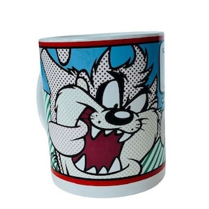 Könnte beinhalten: Eine weiße Keramiktasse mit einem Cartoon-Bild von Taz, dem tasmanischen Teufel aus Looney Tunes. Taz ist mit offenem Mund, bared Zähnen und einem schelmischen Ausdruck dargestellt. Der Hintergrund ist ein farbenfrohes Comic-Design.