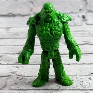 Könnte beinhalten: Eine grüne Kunststoff-Actionfigur mit einem strukturierten, rankenartigen Körper und scharfen roten Augen. Die Figur hat breite Schultern, muskulöse Arme und einen stabilen Stand, mit einer detaillierten Oberfläche, die verwachsene Pflanzen imitiert.
