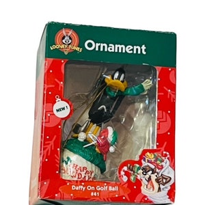 May include: A green, red, and white Christmas ornament box featuring Daffy Duck dressed in a green sweater and a red Santa hat. The ornament is titled "Daffy On Golf Ball #41" and features the Looney Tunes logo.