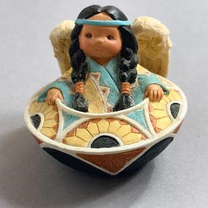 May include: A figurine of a Native American girl with black braids, a blue headband, and angel wings. She is seated in a decorative bowl with a geometric pattern in yellow, blue, and orange. The bowl has a black base.