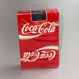 Puede incluir: Una baraja de cartas roja de Coca-Cola. El logotipo de Coca-Cola está en escritura blanca en la parte delantera de la caja. La parte superior de la caja tiene un colgador de plástico transparente.