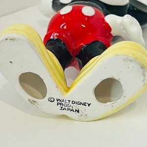 May include: A ceramic figurine of Mickey Mouse standing with his arms outstretched. He is wearing his signature red shorts with black buttons and white circles, black shoes, and white gloves. The figurine is yellow on the bottom and has the text "WALT DISNEY PROD. JAPAN" on the bottom.
