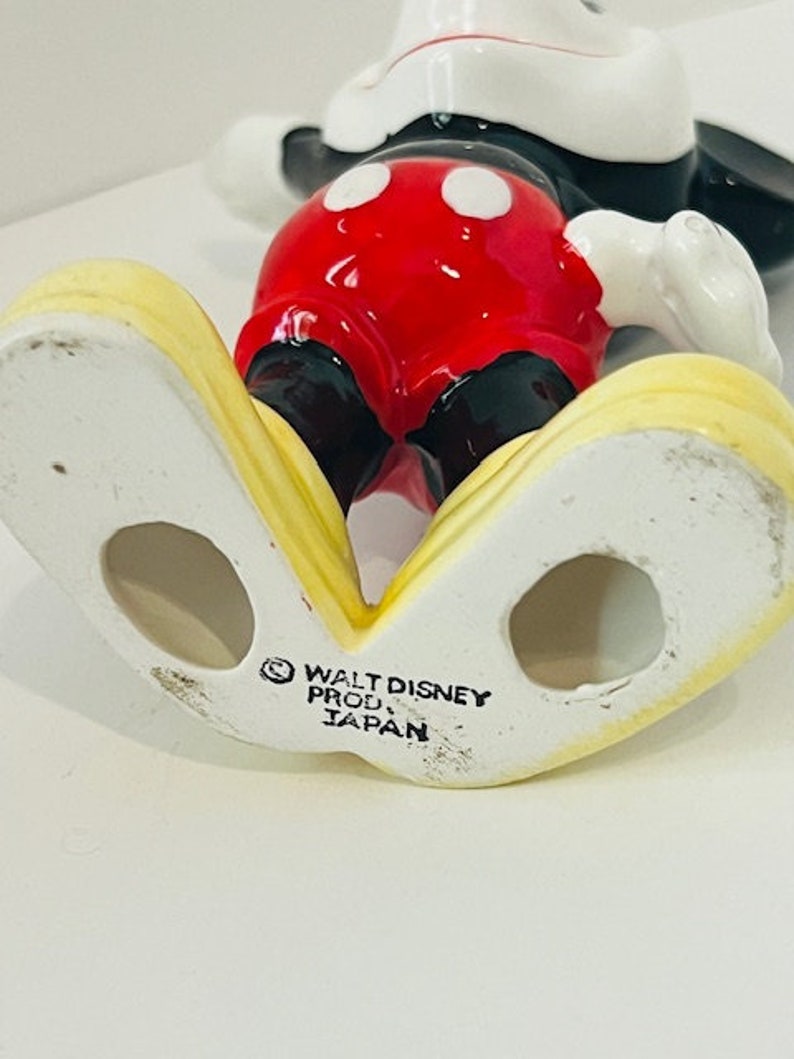 May include: A ceramic figurine of Mickey Mouse standing with his arms outstretched. He is wearing his signature red shorts with black buttons and white circles, black shoes, and white gloves. The figurine is yellow on the bottom and has the text "WALT DISNEY PROD. JAPAN" on the bottom.