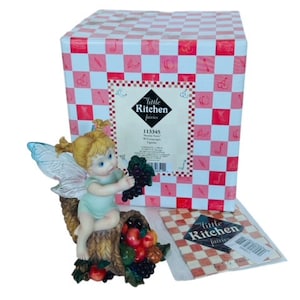 May include: A figurine of a fairy with wings, sitting on a cornucopia filled with fruit and grapes. The fairy is wearing a light blue dress and has blonde hair. The figurine is part of the "Little Kitchen Fairies" collection.