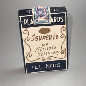 Puede incluir: Una baraja de cartas vintage en una caja. La caja es principalmente azul marino y crema, con las palabras "PLAY CARDS" e "ILLINOIS" en blanco. La caja también presenta el texto "Souvenir of Illinois Tollway" en una fuente decorativa.