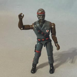May include: A vintage action figure of a soldier in a gray uniform with red accents. The figure has a brown skin tone, a striped headband, and a bandaged face. It is posed with one arm raised.
