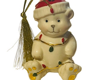 May include: A white porcelain Christmas ornament of a teddy bear wearing a red Santa hat and wrapped in gold and green Christmas lights. The ornament has a gold tassel.