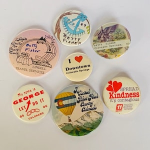 May include: A collection of vintage round pinback buttons with various designs and text. The buttons feature images of a hot air balloon, a house, and text such as "I Downtown Colorado Springs" and "Spread Kindness".