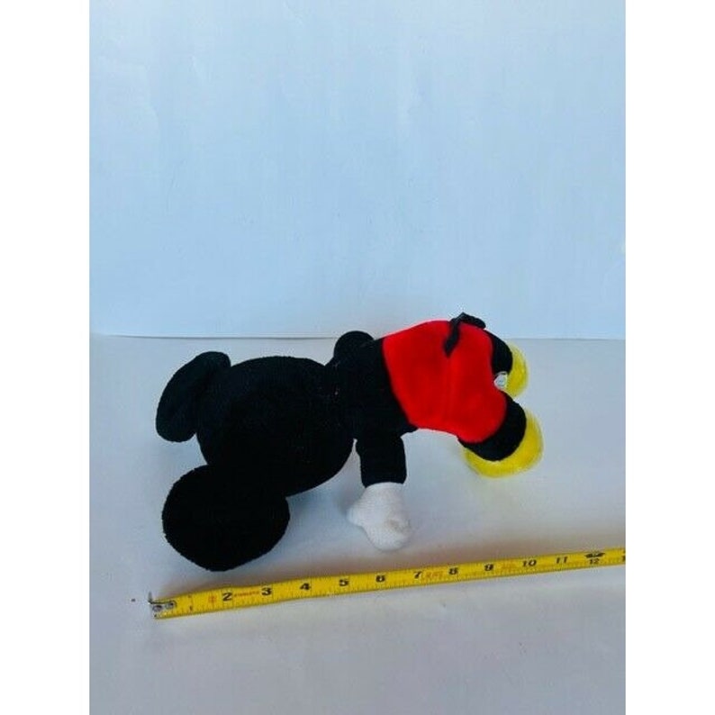 Puede incluir: Un juguete de peluche Mickey Mouse negro y rojo acostado boca abajo. El juguete mide aproximadamente 25 cm de largo.