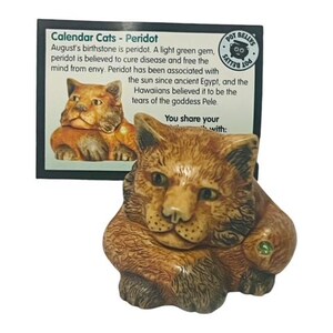 Puede incluir: Una figurita de cerámica de gato marrón y beige con una gema verde en su pata. La figurita está sentada sobre un fondo blanco. El texto en la tarjeta dice "Calendar Cats - Peridot. La piedra natal de agosto es el peridoto. Una gema verde claro, el peridoto se cree que cura enfermedades y libera la mente de la envidia. El peridoto se ha asociado con el sol desde el antiguo Egipto, y los hawaianos creían que eran las lágrimas de la diosa Pele. Compartes tu con:"