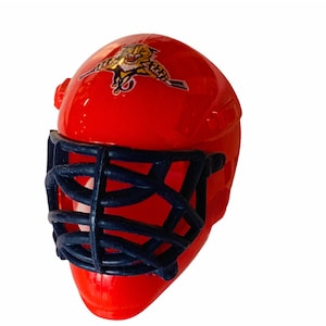 May include: A red hockey goalie mask with a blue cage and a logo of a tiger with a hockey stick. The logo is in orange, yellow, and black.