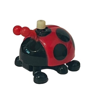 May include: A red and black ladybug wind-up toy with a white winder on top. The toy has six legs and a black and red spotted shell.