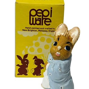 May include: A hand-painted ceramic figurine of a brown rabbit wearing a blue robe with white flowers. The rabbit is holding a white bag in its right paw. The figurine is on a yellow box with the text "pepi ware" and "Hand painted and crafted in, New Brighton, Wallasey, England".