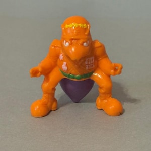 Könnte beinhalten: Orange Kunststoff-Actionfigur einer vogelartigen Kreatur mit lila Unterkörper. Die Figur hat eine gelbe Krone, weiße Augen und einen grün-weißen Gürtel. Die Figur befindet sich in einer hockenden Position.