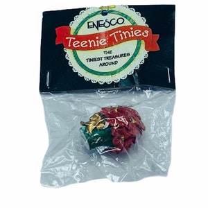 May include: A red poinsettia plant in a green pot with a gold bow. The plant is packaged in a clear plastic bag with a black and white card that reads "Enesco Teenie Tiniest Treasures Around".