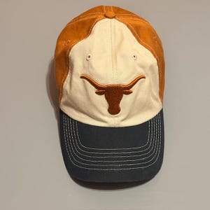 May include: A baseball cap with a burnt orange crown and a navy blue brim. The front panel is off-white with an embroidered longhorn logo in brown. The cap has a curved brim and a button on top.