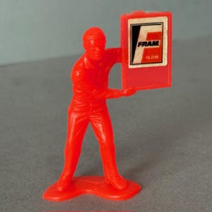 Pode incluir: Pequena figura de plástico vermelho sólido de uma pessoa segurando uma placa retangular vermelha. A placa tem a palavra "FRAM" em letras brancas com um "F" preto e a palavra "FILTERS" em letras brancas.