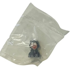 May include: A small black plastic figurine of a cartoon cat with a red nose and white eyes. The figurine is sitting on a clear plastic bag.