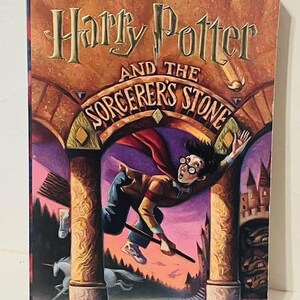 May include: A book cover for "Harry Potter and the Philosopher's Stone" by J.K. Rowling. The cover features an illustration of Harry Potter flying on a broomstick with a purple and pink sky in the background.
