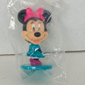 May include: A small plastic figurine of Minnie Mouse wearing a pink bow and a teal dress. She is standing on a teal base.