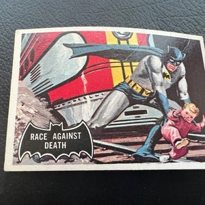 Könnte beinhalten: Eine Vintage-Comic-Karte mit Batman in einem blau-grauen Kostüm, der ein Kind vor einem fahrenden Zug rettet. Die Karte trägt den Text "Race Against Death" in einem schwarzen Fledermaus-Logo.