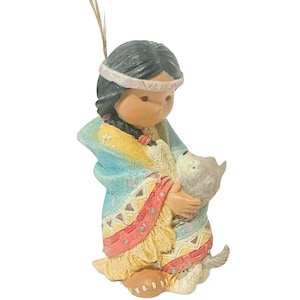 May include: A figurine of a Native American girl wearing a blue and yellow patterned dress and a feathered headdress, holding a small gray and white dog.