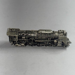 Op de afbeelding: Gedetailleerd miniatuurmodel van een zilverkleurige stoomlocomotief. Het model heeft ingewikkelde details van de motor, waaronder de wielen, schoorsteen en diverse mechanische componenten. De trein staat tegen een neutrale grijze achtergrond.