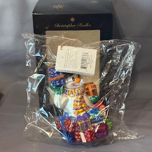Puede incluir: Un adorno de Navidad de vidrio con un muñeco de nieve que lleva una bufanda morada y amarilla y sostiene una pila de regalos coloridos. El adorno está empaquetado en una bolsa de plástico transparente con una etiqueta de Christopher Radko.