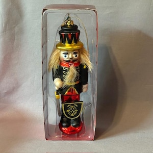 May include: A decorative Christmas ornament shaped like a nutcracker soldier. The ornament is in a clear plastic package. The nutcracker has a black and gold hat, a black uniform with gold accents, and a red base. The ornament is approximately 6 inches tall.