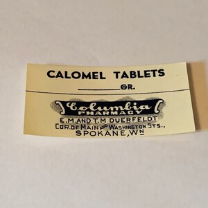 May include: A vintage, cream-colored label for "CALOMEL TABLETS" with black text and a decorative banner for "Columbia Pharmacy." The label includes the address: E.M. and T.M. Duerfeldt, Spokane, WN.