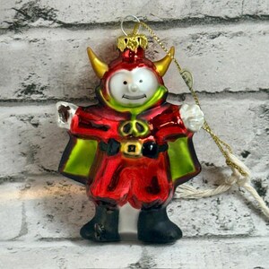 May include: A glass Christmas ornament shaped like a devil character with a white face, yellow horns, and a red suit with a green cape. The figure has black boots, a gold belt buckle, and a gold metal cap with a gold string attached for hanging.