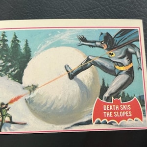 Könnte beinhalten: Eine Vintage-Comic-Sammelkarte mit Batman, der einen schneebedeckten Hang hinunterskifährt. Die Karte hat einen roten und weißen Rand mit dem Text "Death Skis the Slopes" in einem weißen Banner.