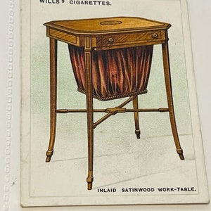 May include: A vintage card featuring a wood work table with a satinwood inlay. The table has a drawer and a fabric-lined basket. The card reads "WILLS'S CIGARETTES" and "INLAID SATINWOOD WORK-TABLE."