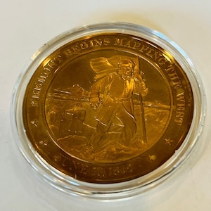 May include: A gold-colored coin with a raised image of a man walking with a staff and a flag. The text around the image reads "A. Fremont Begins Mapping the West June 10, 1842".