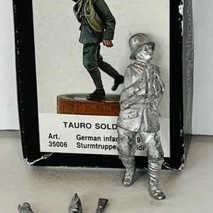May include: A metal model of a German soldier in a white uniform with a helmet and a rifle. The model is standing on a brown base. The box the model is in says "Tauro Sold" and "German Infanterie Sturmtruppen".