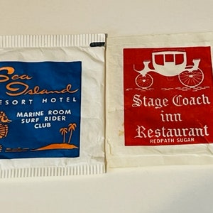 May include: Two vintage sugar packets. One features "Sea Island Resort Hotel" with a seahorse and palm trees on a blue background. The other displays "Stage Coach Inn Restaurant" with a stagecoach illustration on a red background.