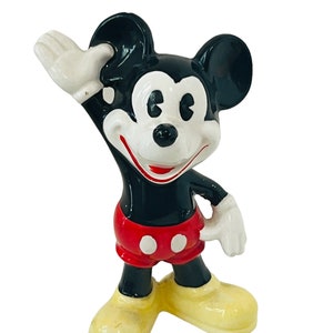 May include: A black and white ceramic figurine of Mickey Mouse. He is wearing red shorts with white buttons and yellow shoes. He is waving with his right hand.