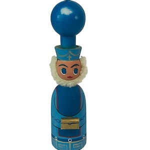May include: A blue wooden candle holder shaped like a person with a blue candle on top. The person is wearing a blue robe with gold trim and a white fur collar. The person is holding a small gold box.