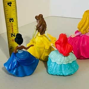 May include: Four plastic Disney princess dolls with different coloured dresses. The dolls are standing on a white surface. The dolls are all facing the same direction. The dolls are all wearing different coloured dresses. The dolls are all wearing different coloured dresses. The dolls are all wearing different coloured dresses.