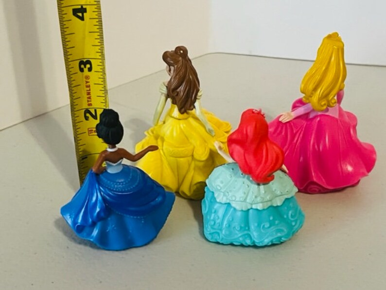 May include: Four plastic Disney princess dolls with different coloured dresses. The dolls are standing on a white surface. The dolls are all facing the same direction. The dolls are all wearing different coloured dresses. The dolls are all wearing different coloured dresses. The dolls are all wearing different coloured dresses.