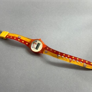 May include: A vintage digital watch with a red and yellow band featuring star and crown patterns. The watch face is orange with a small character illustration and a digital display. The band measures approximately 9 inches in length.